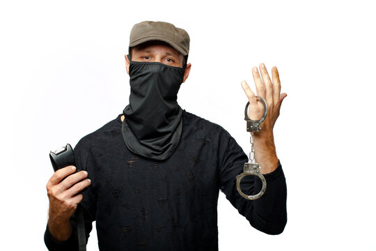 Man in black mask holding handcuffs and Electroshock on white background. The concept of robbery and mafia. Stun gun, taser, electroshock protection against bandits, device for self-defense. 