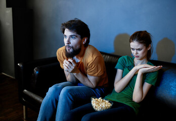 A woman and a man with popcorn on a leather sofa indoors watching TV in the evening