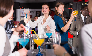 Cheerful girls with coworkers funning in bar during firms party, dancing and toasting drinks