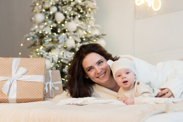 Obraz na płótnie Canvas Mom and son are lying on bed in bright bedroom and are smiling happily. Woman hugs kid boy tightly. The concept of a Christmas morning, home comfort, scandy style. Merry Christmas and Happy New Year.