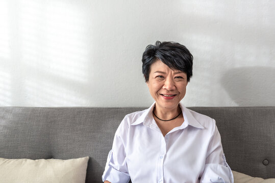 Smile And Happy Middle-aged Gray-haired Asian Woman Wearing White Shirt Pose At Home On A Sunny Morning, A Single Retired Woman Sitting On The Sofa In The Living Room. Lonely Elder Concept