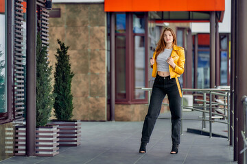 A beautiful redhead woman with freckles in black jeans holds on to the lapels of a yellow jacket near a clothing store.