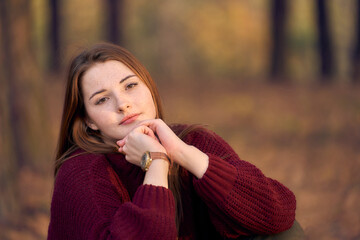 Young woman in a red sweater is sad in the autumn forest. cute face with freckles. Mechanical wrist watch. Autumn nature