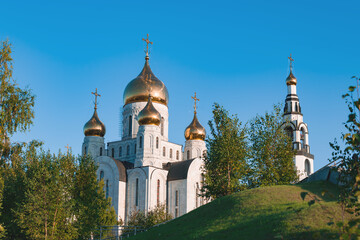 Fototapeta na wymiar Orthodox Church made of white stone with Golden domes among green trees against a blue sky. The topic is faith, religion