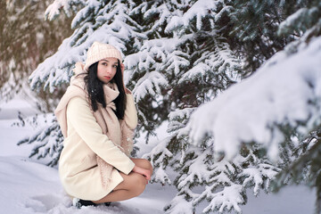 The charming woman squatted down by the snow-covered trees. Fluffy snow lies on fragile spruce branches.