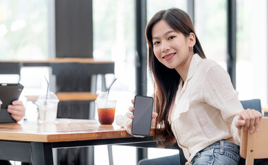 Young casual asian woman showing blank screen smartphone while sitting at the table in coffee shop.