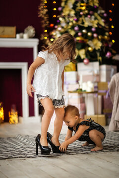 Sibling girls help each other near the Christmas tree in a dark casual interior, children try on their mother's shoes, home photos of children, the concept of new year and christmas