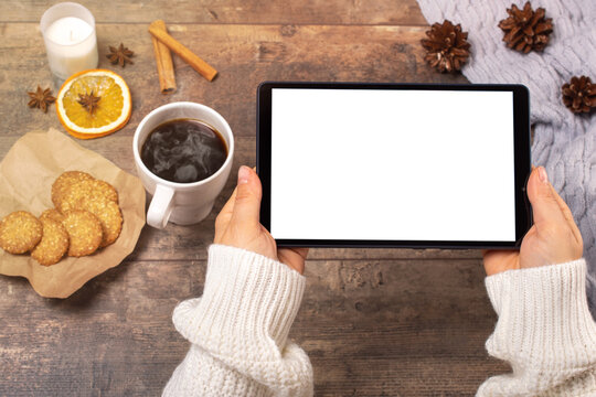 mockup image of woman's hands holding black tablet pc with white blank screen, coffee cup on wooden table. Top view