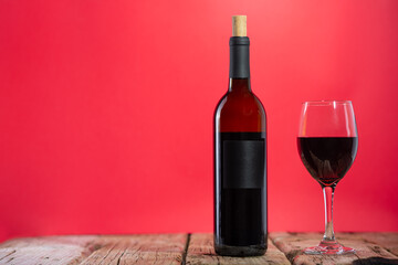Elegant wine bottle and glass with red wine stands on the rustic wooden table on pink background. Space for text. Advertisement concept.