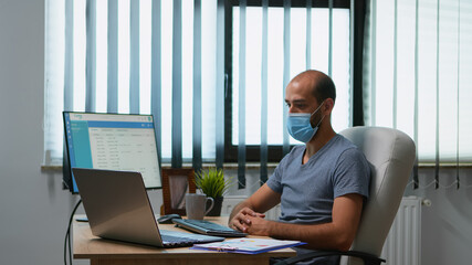 Leader man with medical face mask chatting to colleagues using online video chat during covid-19. Freelancer working in new normal office workplace talking having virtual conference, meeting.