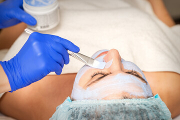 Young woman receiving procedure for applying facial mask by brush in hand of cosmetologist at spa beauty salon
