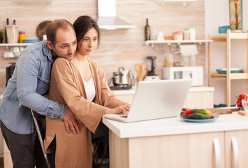 Wife reading on laptop in kitchen while husband is hugging her. Happy loving cheerful romantic in love couple at home using modern wifi wireless internet technology