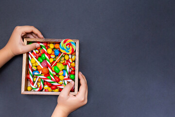 A box of sweets in the hands of a boy on a black background. Various colored candies, lollipops and marmalade in a wooden box, top view, copy space.