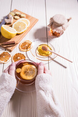 Women's hands in a white knitted sweater hold a Cup of warming tea with lemon, honey and ginger, white background.Close up. Vertical orientation.