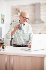 Cheerful senior man during video conference in kitchen on the laptop while enjoying breakfast and a cup of coffee. Elderly person using internet online chat technology video webcam making a video call