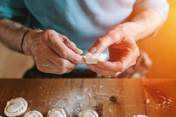 Fototapeta na wymiar hands of senior man cooking and molding small homemade uncooked dumplings with meat on kitchen table. national traditional Russian cuisine. do it yourself. flare