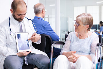 Doctor holding tablet pc with x-ray while explaining diagnosis to disabled elderly woman in wheelchair. Handicapped patient in hospital waiting area. Man in examination room.