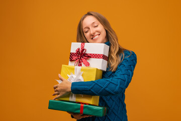 Portrait of a charming young woman in knitted sweater holding stack of present boxes. Studio shot, yellow background