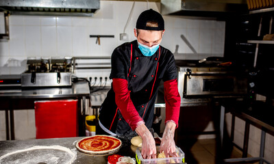 Pizza cooking concept, chef in medical mask puts ingredients on pizza

