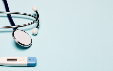 A digital thermometer and a stethoscope lie on a light blue background. Copy space. Medical concept