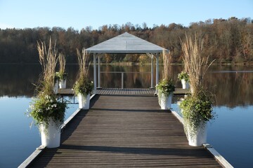 A wooden pier with white gazebo and flower pots with plants over a lake surrounded by forest in...