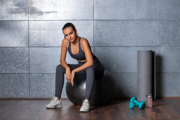Female fitness trainer sits on a gymnastic ball with fitness equipment, dumbbells, mat and a bottle after workout