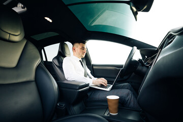 Self-driving electric car autopilot demanding driver attention to hold steering wheel and take control on highway. Businessman working on laptop at sunset.