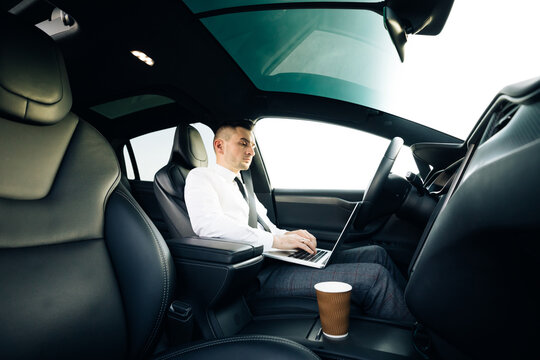 Self-driving electric car autopilot demanding driver attention to hold steering wheel and take control on highway. Businessman working on laptop at sunset.