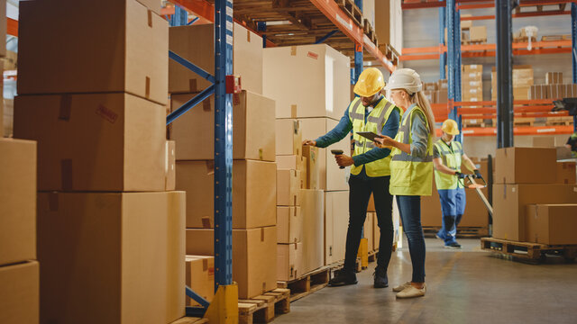 Retail Warehouse full of Shelves with Goods in Cardboard Boxes, Male Worker and Female Supervisor Holding Digital Tablet Discuss Product Delivery while Scanning Packages.Distribution Logistics Center