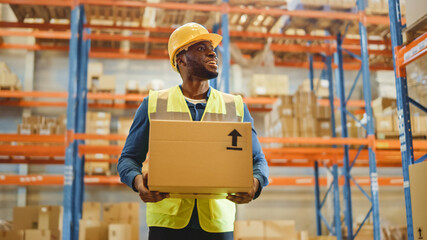 Handsome Male Worker Wearing Hard Hat Holding Cardboard Box Walking Through Retail Warehouse full of Shelves with Goods. Working in Logistics and Distribution Center. Front Shot.