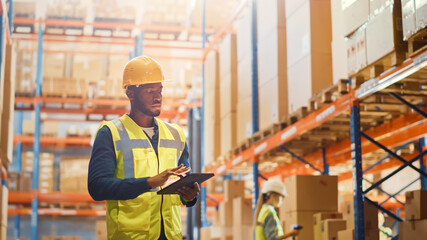 Handsome Male Worker Wearing Hard Hat Checks Products Stock and Inventory with Digital Tablet Computer Standing in Retail Warehouse full of Shelves with Goods. e-Commerce Distribution, Logistics