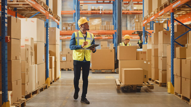 Handsome Male Worker Wearing Hard Hat Holding Digital Tablet Computer Walking Through Retail Warehouse full of Shelves with Goods. Working in Logistics and Distribution Center.