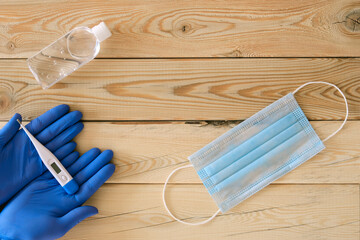two hands in blue gloves hold a white thermometer, a blue mask and an antiseptic in a transparent plastic bottle on a wooden background