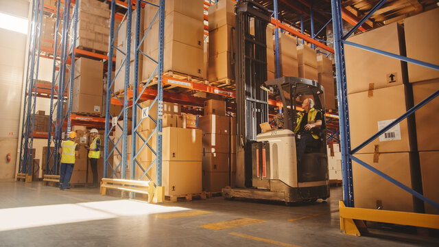 Retail Warehouse full of Shelves with Goods: Electric Forklift Truck Operator Lifts Pallet with Cardboard Box on a Shelf. People Working, Scanning Products, Using Trucks in Logistics Delivery Center