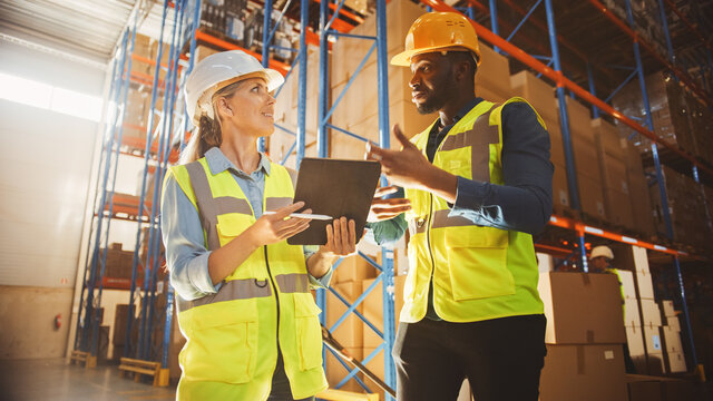 Male and Female Supervisors Holding Digital Tablet Talk about Product Delivery in Retail Warehouse full of Shelves with Goods. Workers in Distribution Logistics Center. Low Angle Shot