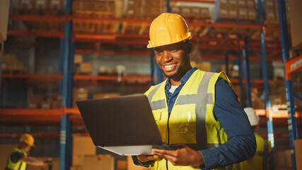 Professional Male Worker Wearing Hard Hat Holding Laptop Computer Checking Stock and Inventory in the Retail Warehouse full of Shelves with Goods and People Working.