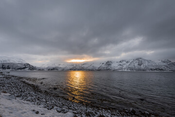 Scenic view over morning sun breaking through dense clouds behind snow-clad mountains near lake on the Lofoten islands archipelago in Norway
