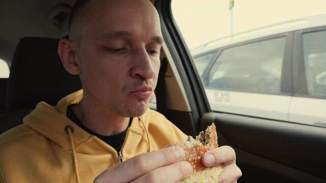 Man driver sit in a car hold eat a big meat burger from mcdrive bite it and chew