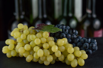 In the foreground there are white and blue grapes, in the background are not clear wine bottles.
