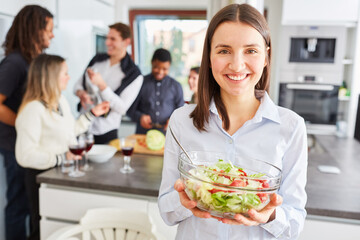 Lächelnde Frau in Küche mit Freunden hält Schüssel  frischen Salat in Händen