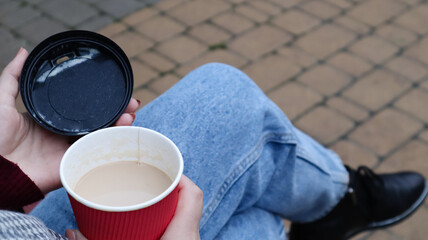 Female hands are holding morning takeaway coffee and closing a paper cup with a lid while sitting on a park bench, close-up of hands.