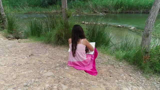 Ni&ntilde;a vestida rosa sentada en la orilla de rio y dron orbitando