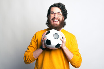 A bearded man with glasses is looking excited at the camera holding a soccer ball with both hands .