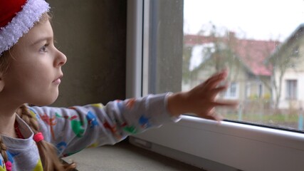 Little girl by the window in Santa hat. Orphans and Christmas . Orphanage life and holidays.