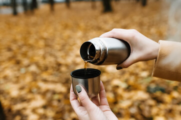 Fototapeta premium Woman pours tea from a thermos, outdoors. Close-up of female hands and a mug with tea. Autumn background.