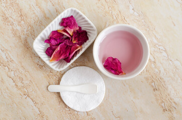 Obraz na płótnie Canvas Two small white bowl with rose water (toner), dry rose petals and cotton pad. Ingredients for preparing homemade cosmetics. Natural beauty treatment recipe, zero waste concept. Top view, copy space