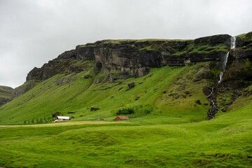 Fototapeta na wymiar Icelandic fresh landscape with green grass, hills and few houses full of waterfalls near a highway