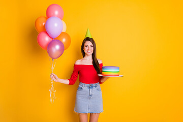 Photo of cute charming cheerful young girl shiny smiling hold many balloons cake birthday concept wear cone shirt open shoulders denim skirt isolated bright yellow color background