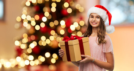 Obraz na płótnie Canvas winter holidays and people concept - happy smiling teenage girl in santa helper hat holding gift box over christmas tree lights background