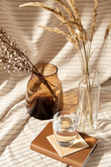 home improvement and decoration concept - still life of water in glass, book, decorative dried flowers in vase and bottle over drapery